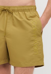 Shorts de bain pour hommes en vert olive, fabriqués dans un tissu lisse. Dotés d'une taille élastique avec cordon de serrage et d'un logo brodé sur le côté.