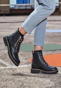 Black leather ankle boots with a side zipper, lace-up front, stud accents, and a rugged sole. Worn with light gray trousers.