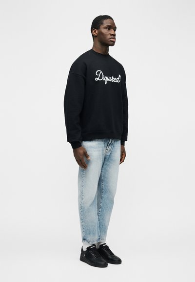 Άνδρας που στέκεται φορώντας μαύρο φούτερ με λευκό κείμενο "Dsquared", ανοιχτόχρωμα μπλε τζιν, λευκές κάλτσες και μαύρα αθλητικά σε απλό φόντο.