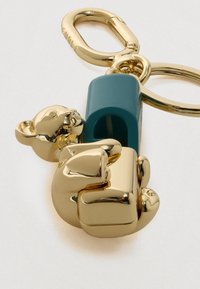 Aranyozott medve bűbáj türkizkék blokkhoz csatlakoztatva, rákcsipesz kapoccsal és kulcskarikával, fényes felülettel és sima textúrával.