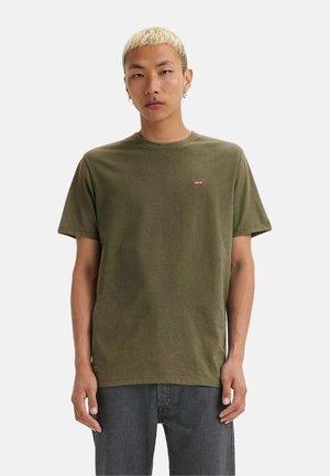 Ung mand med kort, blond hår iført en olivengrøn Levi's t-shirt og mørke jeans, står foran en ensfarvet hvid baggrund.
