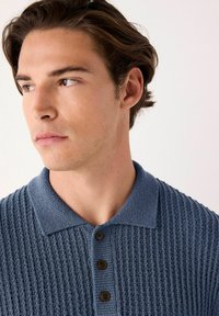 Polo shirt texturată albastră cu un design cu nervuri, guler moale și trei nasturi maro. Materialul pare ușor și respirabil.