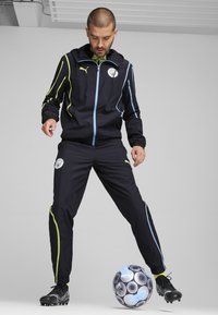 Giacca sportiva nera con accenti blu e lime, chiusura con zip e logo della squadra. Pantaloni neri con vita elasticizzata. Palla da calcio nera ai piedi.