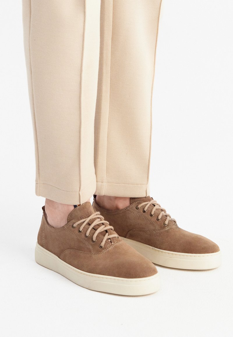 Braune Wildleder-Schnürschuhe mit einer cremefarbenen Gummisohle, abgerundetem Zeh und minimalen Nähdetails, kombiniert mit beigen Jogginghosen.