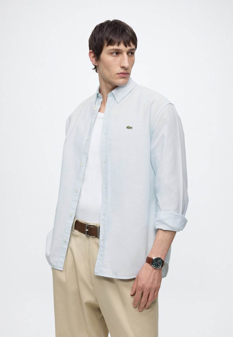 Uomo che indossa una camicia azzurra chiara con bottoni sopra una t-shirt bianca, pantaloni beige, cintura marrone e orologio da polso, guarda di lato su uno sfondo bianco semplice.