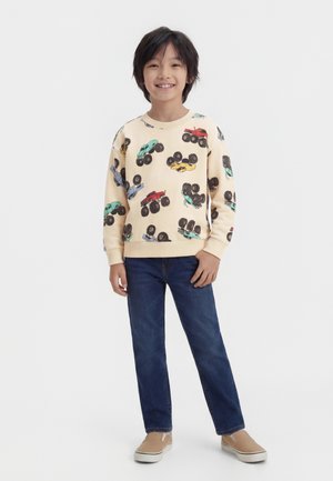 Barn står og smiler, iført creme sweatshirt med farverigt monster truck-print, blå jeans og beige slip-on sko.