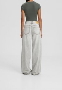 Femme debout, dos à la caméra, portant un haut moulant à manches courtes vert foncé et un jean gris clair à jambes larges sur fond gris.