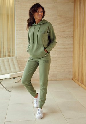 Olive green hoodie and matching joggers, soft fabric, features front pocket and text detail. Worn with white sneakers on tiled floor.