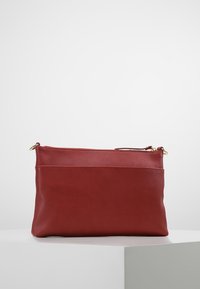 Czerwona skórzana kopertówka z zapięciem i elementami wykończenia w kolorze złotym, wyeksponowana na białej powierzchni na jednolitym tle.