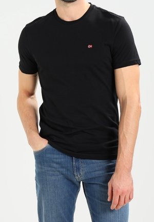 Uomo che indossa una maglietta nera a maniche corte con un piccolo logo rosso e bianco sul petto e jeans blu, con la mano in tasca.