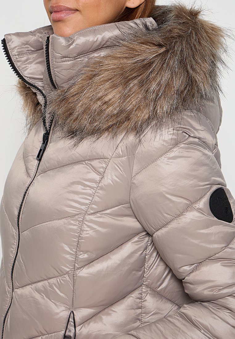 Donna che indossa un giubbotto invernale trapuntato di un beige chiaro con cerniera nera e un ampio bordo in pelliccia sintetica marrone.