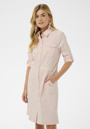 Kobieta o długich blond włosach, ubrana w jasnoróżową sukienkę zapinaną na guziki z podwiniętymi rękawami i różowe kwiatowe kolczyki, prawa ręka w kieszeni.