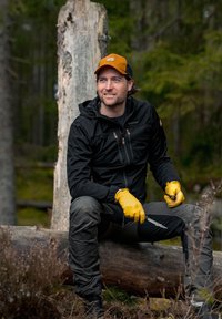 Black zippered jacket with a hood, olive green pants, yellow gloves, sitting on a log in a forest. Notable details include a small knife and wood textures.