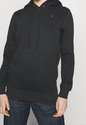 Person trägt einen schwarzen Kapuzenpullover mit Kordelzug und dunkelblaue Jeans vor einfachem Hintergrund.