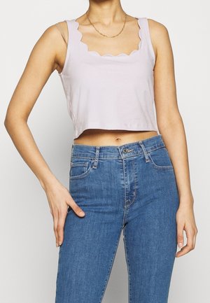 Woman wearing a lavender scalloped-edge crop top and high-waisted blue jeans against a plain background.