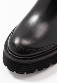 Schwarze Lederstiefel mit runder Zehenpartie, strukturiertem Obermaterial und dickem, geriffelten Sohlen. Verziert mit dekorativen Nähten entlang der oberen Kante.