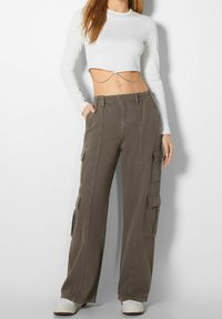 Pantalones cargo en algodón verde oliva con múltiples bolsillos laterales, un corte holgado y costuras visibles. Combinados con una camiseta de manga larga blanca, corta.