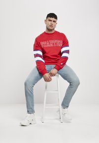 Roter Pullover mit "STARTER"-Aufschrift, kontrastierenden blau-weißen Streifenärmeln, kombiniert mit hellblauen Jeans und weißen Sneakers.