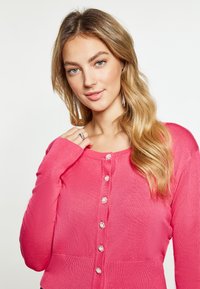 Cardigan rosa brillante con scollo rotondo, maniche lunghe e bottoni decorativi simili a gemme sulla parte anteriore; texture liscia.