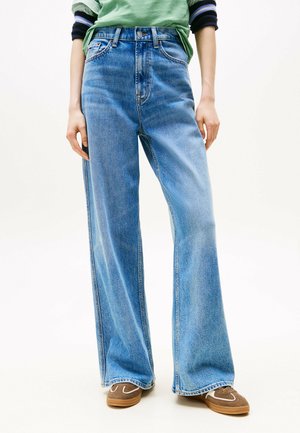 Persona che indossa jeans azzurri a gamba larga, camicia verde, canottiera a righe e sneakers marroni, in piedi contro uno sfondo bianco.