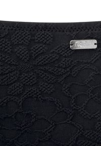 Tissu noir texturé avec un motif floral, ornée d'une étiquette en métal argenté affichant le nom de la marque en haut.