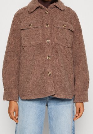 Veste sherpa marron texturée à boutons avec poches poitrine à rabat, portée sur un haut foncé et un jean bleu clair.