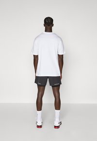T-shirt blanc à manches courtes et shorts de sport noirs. Le T-shirt a un design simple ; les shorts présentent des détails de logo. Personne de dos.