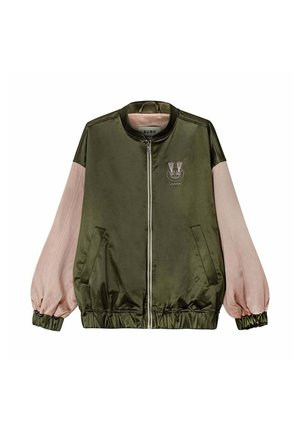 Junge Frau mit gestrickter Kapuzenmütze und zweifarbiger Jacke beißt in ein Gebäck, während sie an einem Sandstrand steht.