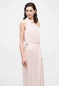 Žena u blijedo ružičastoj, bezrukavnoj haljini na jedno rame s naboranim izrezom i dugom suknjom, stoji na neutralnoj bijeloj pozadini.