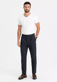 Biała koszulka V-neck, ciemne spodnie z zakładkami, brązowe skórzane buty. Model stoi na jednolitym tle, prezentując swobodny, ale dopasowany wygląd.