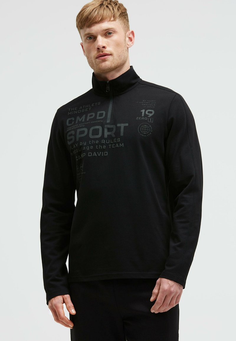 Schwarzes Sportoberteil mit langen Ärmeln und hohem Kragen, ausgestattet mit einem Teilreißverschluss und bedrucktem Text in verschiedenen Grautönen für Designdetails.