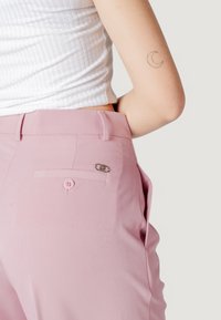 Pantaloni rosa su misura con una texture liscia, dotati di una tasca posteriore con bottone e un accento argentato. Indossati con un top bianco a costine.