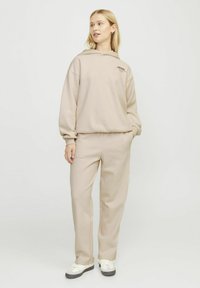 Sudadera beige y pantalones de chándal a juego, fabricados con un tejido suave. La sudadera cuenta con un bolsillo frontal y texto con logo, con un corte relajado. Zapatillas blancas.