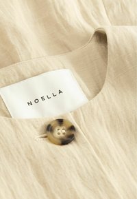 Tissu beige avec un gros plan sur un bouton écaille de tortue et une étiquette de vêtement blanche portant l'inscription « NOELLA » cousue à l'intérieur du col.