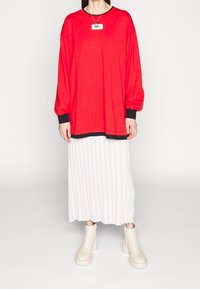 Röd överdimensionerad långärmad skjorta med svarta manschetter, ihop med en vit veckad kjol och beige anklarstövlar med klumpiga klackar.