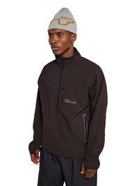 Homme portant un bonnet gris et une veste noire à fermeture éclair partielle avec poches zippées sur la poitrine et les côtés, debout les mains détendues le long du corps.