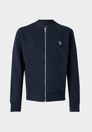 Jeune homme debout, les mains dans les poches, portant une veste bleu marine, un t-shirt blanc, un jean bleu et des chaussures marron sur un fond uni.