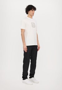 White cotton t-shirt with a square logo in a beige color on the chest, paired with dark denim jeans and white sneakers.