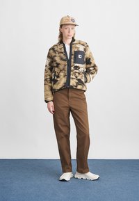 Tan and black fuzzy jacket with a front zipper, black patch pocket, paired with brown pants and white sneakers on a blue carpet backdrop.