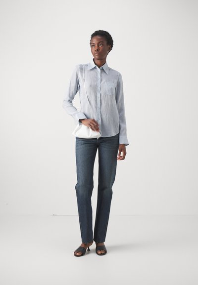 Lichtblauw, getextureerd overhemd met knopen en een borstzak, gecombineerd met donkerdenim jeans en zwarte open-teen sandalen, terwijl een kleine witte tas wordt vastgehouden.