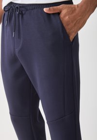 Close-up of a person wearing navy blue sweatpants with a drawstring waist and hand placed inside the right pocket.