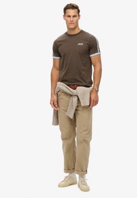 Camiseta de algodón marrón con cuello y acentos de manga en azul claro, combinada con pantalones beige y un suéter de punto beige envuelto. Zapatillas claras.
