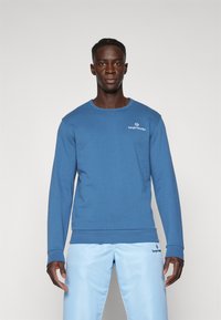 Sudadera azul con mangas largas y escote redondo. Presenta un pequeño logo blanco en el pecho. Textura suave, corte casual.