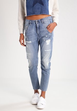 Modelka ubrana w jasnoniebieskie jeansy z przetarciami, krótki sweter z niebieską gwiazdą oraz białe tenisówki wsuwane, stoi z jedną ręką w kieszeni.