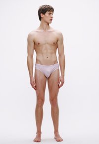 Meesmodell heleroosades ujumispükstes, istuva lõikega, siledapinnaline, minimaalne vöökoht, seisab sirgelt tavalise valge tausta ees.