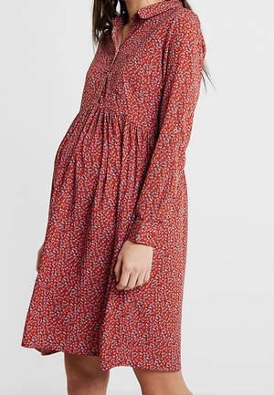 Femme portant une robe rouge ample à manches longues avec un petit motif floral blanc et un col, debout devant un fond uni.