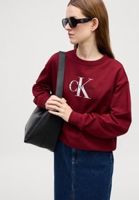 Maglione bordeaux con logo Calvin Klein, abbinato a una borsa tote nera e una gonna di jeans scuri. Occhiali da sole oversize neri completano il look.