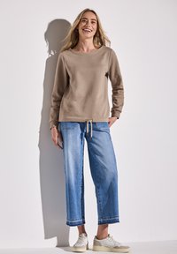 Beige mikina s kulatým výstřihem, spárovaná s světle modrými džínami s širokými nohavicemi a třásněmi na lemu a šňůrkou v pase. Bílé tenisky.