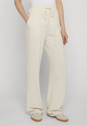 Beige locker sitzende Hose mit elastischem Bund und Kordelzug, getragen mit beigen und blauen flachen Schuhen, Hände in den Taschen, vor einem schlichten Hintergrund.