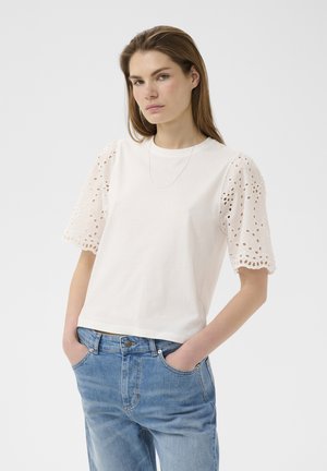 Egyenes hajú nő fehér felsőt visel, amelyen szembefűzött pöttyös pufó ujj található, és kék farmert, zsebekbe dugott kézzel, fehér háttér előtt áll.